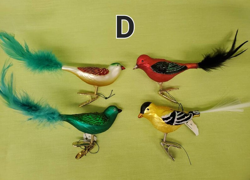 May include: Four glass bird ornaments with feather tails. The birds are red, green, white, and yellow with black accents. The ornaments have a clip on the bottom for hanging.