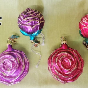 May include: Four glass Christmas ornaments shaped like roses. Two are pink with gold glitter, one is purple with gold glitter, and one is teal with gold glitter.