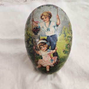 Könnte beinhalten: Ein vintage Osterei mit einer Papiermaché-Außenseite. Das Ei zeigt eine farbenfrohe Illustration von zwei Kindern auf einer Schaukel in einem Garten.