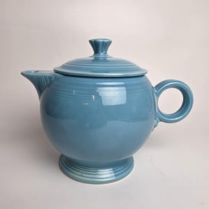 May include: A light blue ceramic teapot with a lid and handle. The teapot has a round body and sits on a pedestal base. The handle is a simple loop, and the lid has a small knob. The teapot is a solid, glossy color.