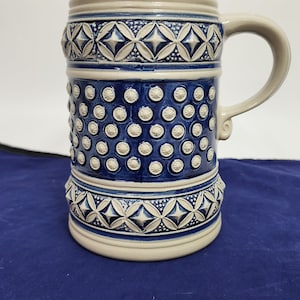 May include: A ceramic beer stein with a handle. The stein is primarily blue and beige, with intricate embossed patterns. The design includes geometric shapes and small circular accents. The handle is curved and the stein has a lid.