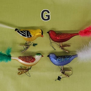 May include: Four glass bird ornaments with feathered tails. The birds are yellow with black stripes, red, blue, and white with brown stripes. The ornaments are hanging from gold clips.