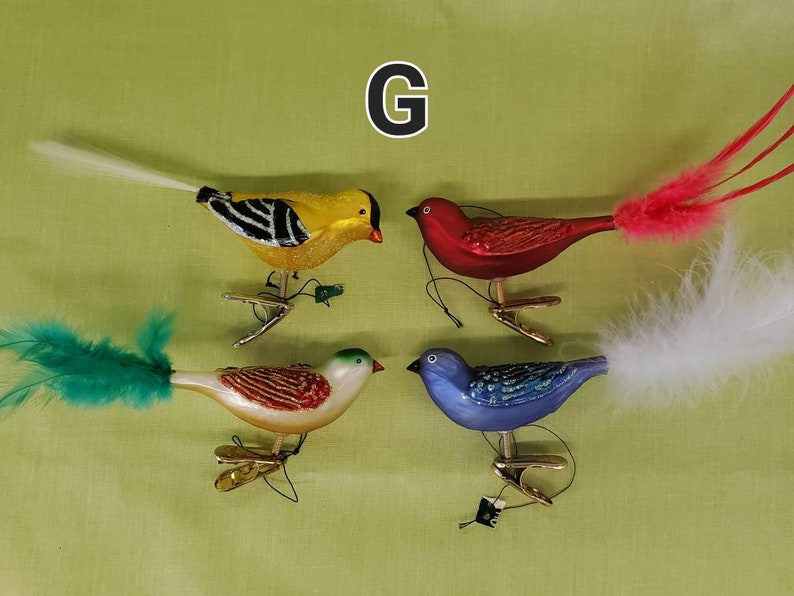 May include: Four glass bird ornaments with feathered tails. The birds are yellow with black stripes, red, blue, and white with brown stripes. The ornaments are hanging from gold clips.