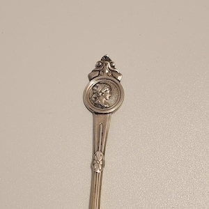 May include: Silver spoon with a decorative handle featuring a round medallion with a profile of a woman's face. The handle is ornate and has a floral design at the bottom.