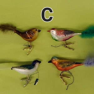 May include: Four glass bird ornaments with feathers. The birds are brown, pink, blue, and orange. Each bird has a gold clip on the bottom.