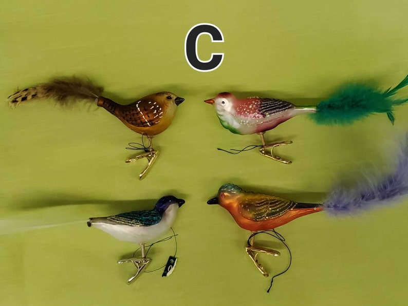 May include: Four glass bird ornaments with feathers. The birds are brown, pink, blue, and orange. Each bird has a gold clip on the bottom.