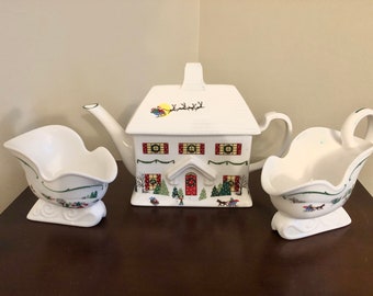May include: A white ceramic teapot shaped like a house with a Christmas scene on the front. The teapot is accompanied by two matching creamer and sugar bowls shaped like sleighs.