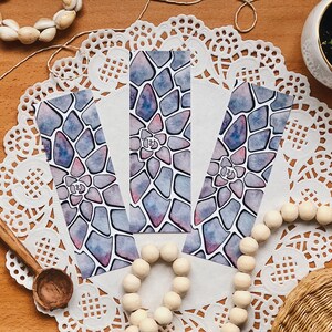 May include: Three watercolour bookmarks with a succulent design in blue, purple, and pink hues. The bookmarks have a geometric pattern and a white background. The image also includes decorative elements like a wooden spoon, beads, and a small succulent plant.