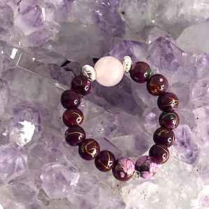 Peut inclure: Un bracelet en perles violettes et dorées avec un charme en pierre rose. Le bracelet repose sur un lit de cristaux d'améthyste.