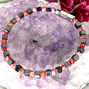 May include: A beaded bracelet featuring a mix of square, dark gray hematite beads and round, patterned beads in shades of red, orange, and white. The bracelet is accented with a silver magnetic clasp. The background includes a purple crystal and pink flowers.