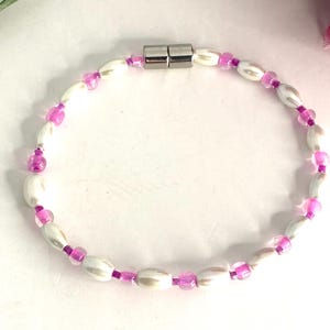 May include: A white and pink beaded bracelet with a magnetic clasp. The beads are oval shaped and have a slight iridescent sheen.