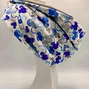 Puede incluir: Un scrunchie de tela blanca con un estampado de Mickey Mouse azul y plateado con purpurina. El scrunchie est&aacute; en una cabeza de maniqu&iacute; blanca.