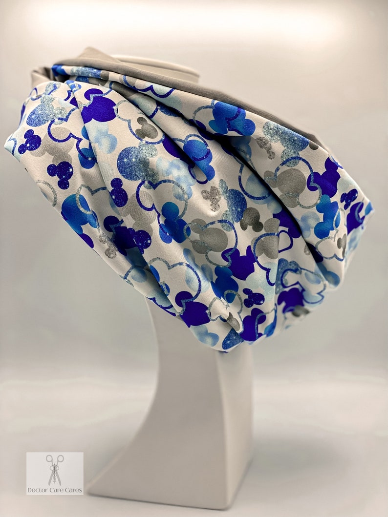 Puede incluir: Un scrunchie de tela blanca con un estampado de Mickey Mouse azul y plateado con purpurina. El scrunchie est&aacute; en una cabeza de maniqu&iacute; blanca.