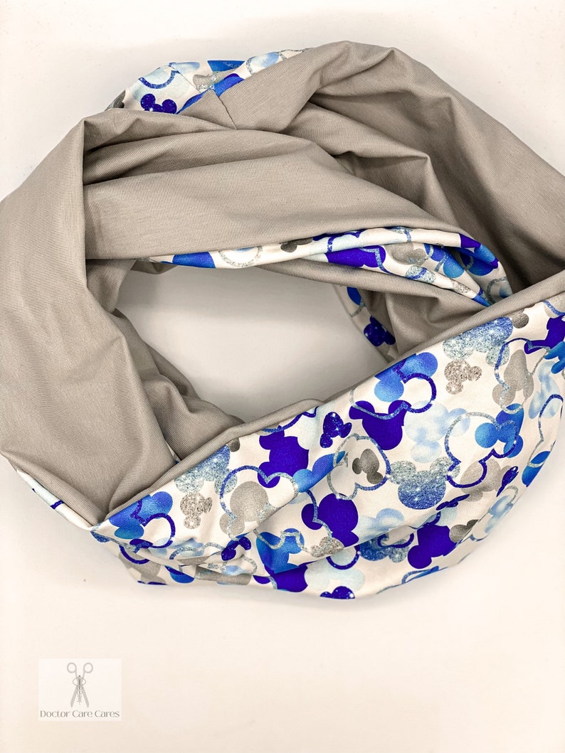 Puede incluir: Una bufanda infinita reversible con un lado gris y un lado blanco con un estampado de Mickey Mouse azul y plateado.