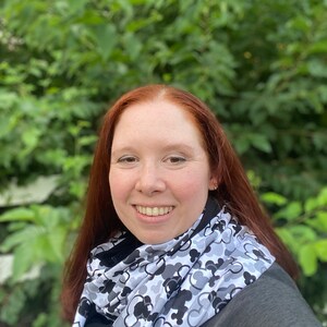 May include: A woman with long, red hair is wearing a black jacket and a white scarf with a black and gray Mickey Mouse pattern. She is smiling and looking at the camera.
