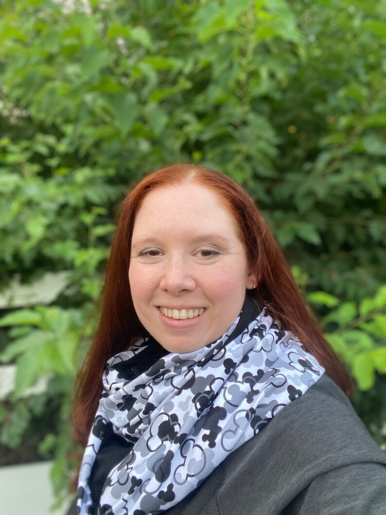 May include: A woman with long, red hair is wearing a black jacket and a white scarf with a black and gray Mickey Mouse pattern. She is smiling and looking at the camera.