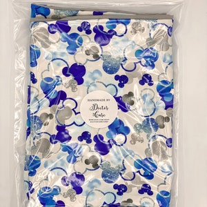 Puede incluir: Una tela blanca con un patr&oacute;n de Mickey Mouse azul y plateado con purpurina. La tela est&aacute; empaquetada en una bolsa de pl&aacute;stico transparente. La bolsa tiene una etiqueta que dice "Handmade by Doctor Care" e incluye una direcci&oacute;n de sitio web.
