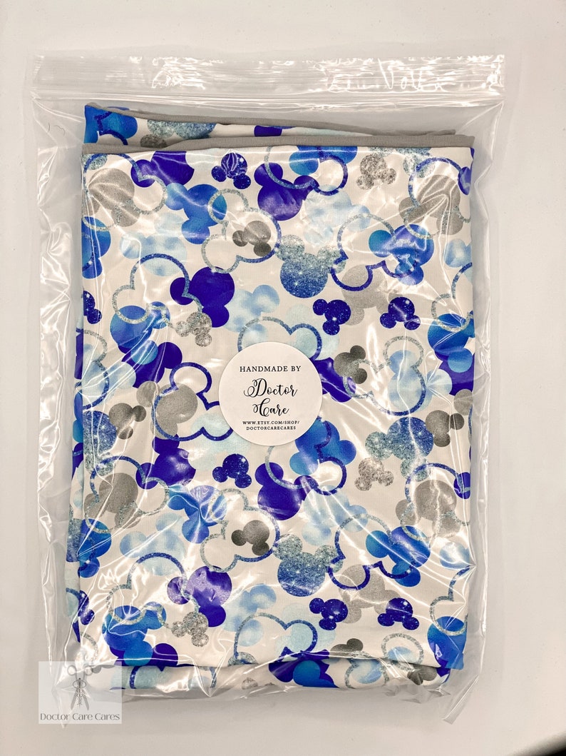 Puede incluir: Una tela blanca con un patr&oacute;n de Mickey Mouse azul y plateado con purpurina. La tela est&aacute; empaquetada en una bolsa de pl&aacute;stico transparente. La bolsa tiene una etiqueta que dice "Handmade by Doctor Care" e incluye una direcci&oacute;n de sitio web.