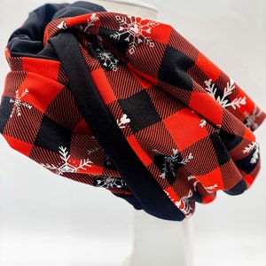 May include: A red and black plaid infinity scarf with white snowflake designs. The scarf is folded and displayed on a white mannequin.