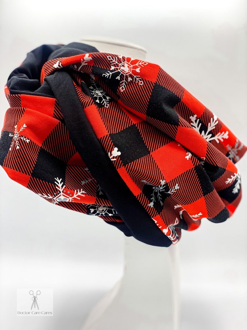 May include: A red and black plaid infinity scarf with white snowflake designs. The scarf is folded and displayed on a white mannequin.