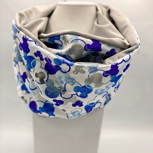 Puede incluir: Una bufanda infinita blanca con un estampado de Mickey Mouse azul y plateado. La bufanda es reversible con un lado gris.