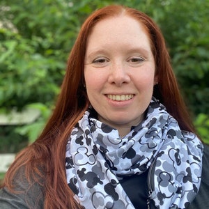 May include: A woman with long red hair is wearing a gray and black infinity scarf with a zipper. The scarf has a pattern of Mickey Mouse heads.