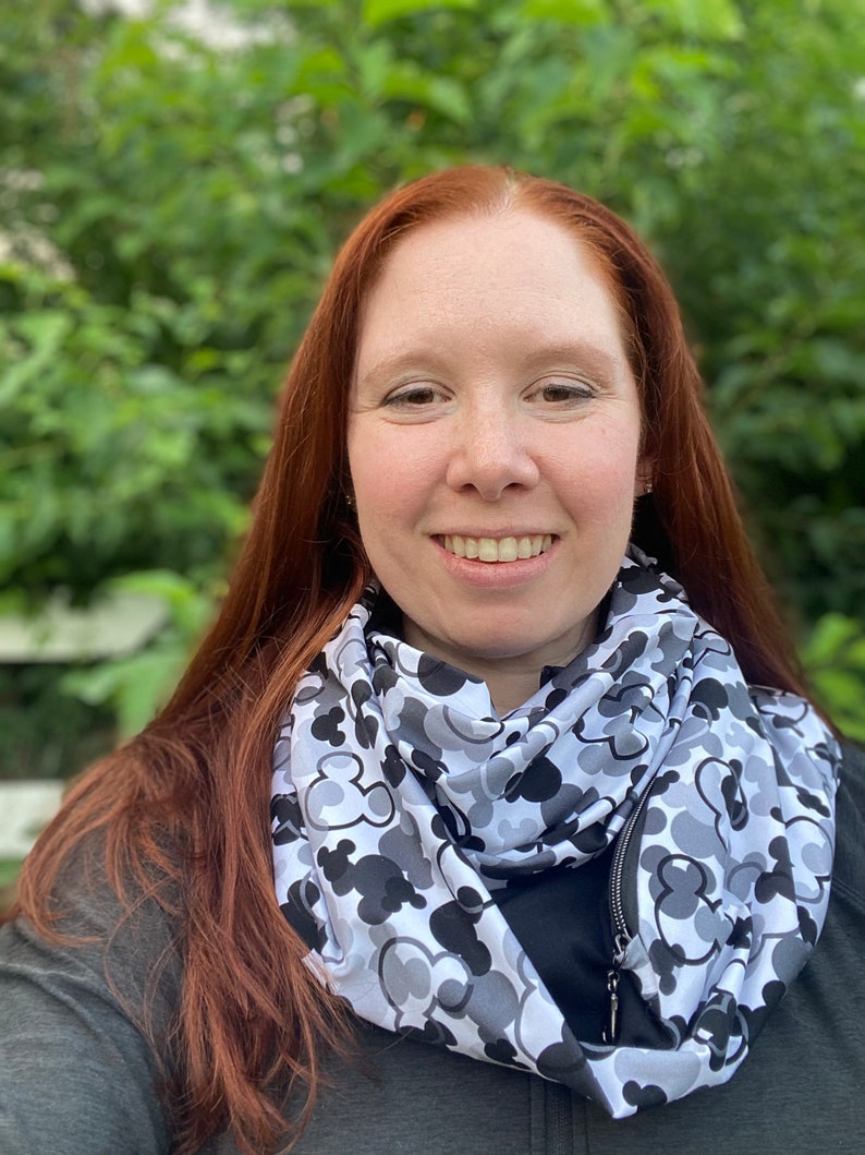 May include: A woman with long red hair is wearing a gray and black infinity scarf with a zipper. The scarf has a pattern of Mickey Mouse heads.