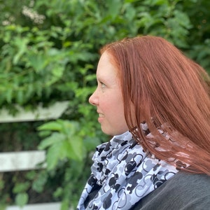 May include: A woman with long red hair is wearing a white and black infinity scarf with a repeating pattern of Mickey Mouse heads. The scarf is worn around her neck and is visible against a green leafy background.