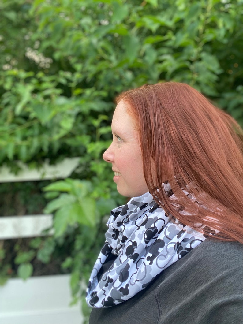 May include: A woman with long red hair is wearing a white and black infinity scarf with a repeating pattern of Mickey Mouse heads. The scarf is worn around her neck and is visible against a green leafy background.