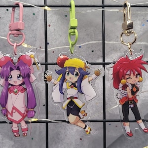 Op de afbeelding: Drie anime-personage sleutelhangers met verschillende haarkleuren en outfits. De sleutelhanger links heeft paars haar en een roze en witte outfit. De sleutelhanger in het midden heeft blauw haar en een gele en witte outfit. De sleutelhanger rechts heeft rood haar en een rode en zwarte outfit.