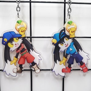 Może przedstawiać: Dwa przezroczyste breloki akrylowe z postacią z kreskówki o czarnym futrze, białych uszach i niebieskim kapeluszu. Postać trzyma złoty pierścień z zielonym klejnotem. Jeden brelok przedstawia postać w czerwonej koszuli i niebieskich spodniach, a drugi brelok przedstawia postać w niebieskiej koszuli i niebieskich spodniach.