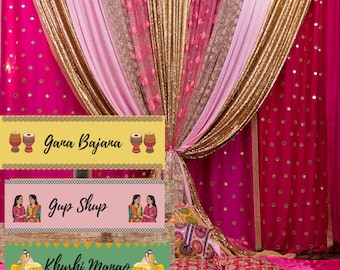 May include: A vibrant scene with a pink and gold backdrop, featuring three signs with text and illustrations. The signs read "Gana Bajara," "Gup Shup," and "Khushi Manao." Decorative pillows are scattered on a pink surface.