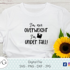 May include: White t-shirt with black text that reads "I'm not overweight I'm under tall!" with a scale icon. The shirt is lying on a white wooden surface with a sunflower in the background.