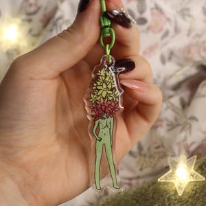May include: A clear acrylic keychain featuring a green figure with a floral head of yellow and red flowers. The keychain is attached to a green clasp. The figure is standing with one hand on its hip.