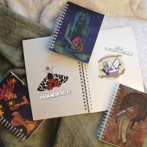 May include: Four spiral bound notebooks with different designs. The first notebook has a blue and green illustration of a woman holding a smaller figure. The second notebook has a black and white illustration of a moth with the text "From Neon Decay". The third notebook has a black and white illustration of a person sitting on a toilet with the text "From The Self-Made Man". The fourth notebook has a brown and black illustration of a person with a black and white design on their clothing.