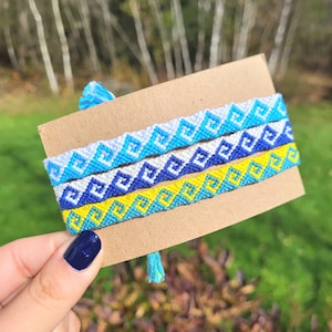 May include: Three friendship bracelets with a geometric pattern in shades of blue, yellow, and white. The bracelets are displayed on a brown card.