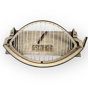 May include: A wooden model of the Gateshead Millennium Bridge. The model is made of light-coloured wood with a curved design and vertical support cables. The bridge's name is printed on the wooden base.