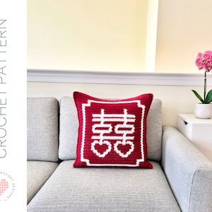 May include: A red crochet pillow with a white double happiness symbol and heart design. The pillow is square-shaped and has a white border. The words "CROCHET PATTERN" are printed on the left side of the image.