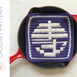 May include: Purple and white crocheted pot holder with a geometric design. The pot holder is sitting in a red cast iron skillet.