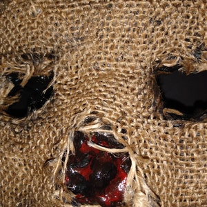 Puede incluir: Primer plano de una m&aacute;scara de arpillera con grandes agujeros para los ojos y una boca abierta. El &aacute;rea de la boca est&aacute; manchada con pintura roja y negra, lo que sugiere una apariencia sangrienta o macabra. La m&aacute;scara tiene una superficie rugosa y texturizada.