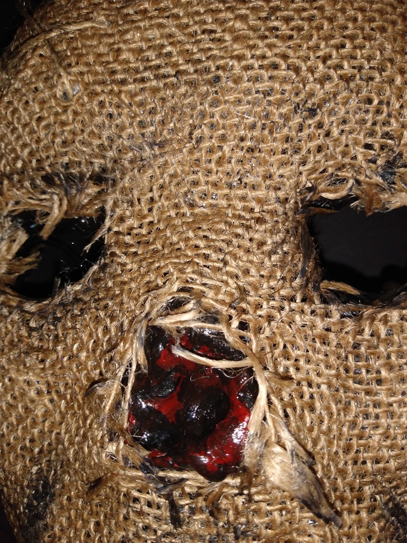 Puede incluir: Primer plano de una m&aacute;scara de arpillera con grandes agujeros para los ojos y una boca abierta. El &aacute;rea de la boca est&aacute; manchada con pintura roja y negra, lo que sugiere una apariencia sangrienta o macabra. La m&aacute;scara tiene una superficie rugosa y texturizada.