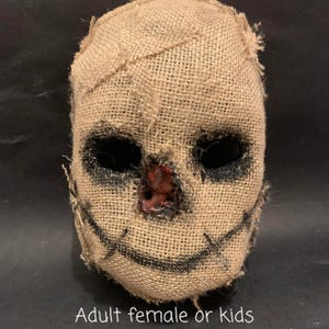 Puede incluir: Una m&aacute;scara de espantap&aacute;jaros de arpillera con costuras, agujeros negros para los ojos y una sonrisa cosida. La nariz est&aacute; pintada de rojo. El texto "Adult female or kids" est&aacute; en la parte inferior de la imagen.