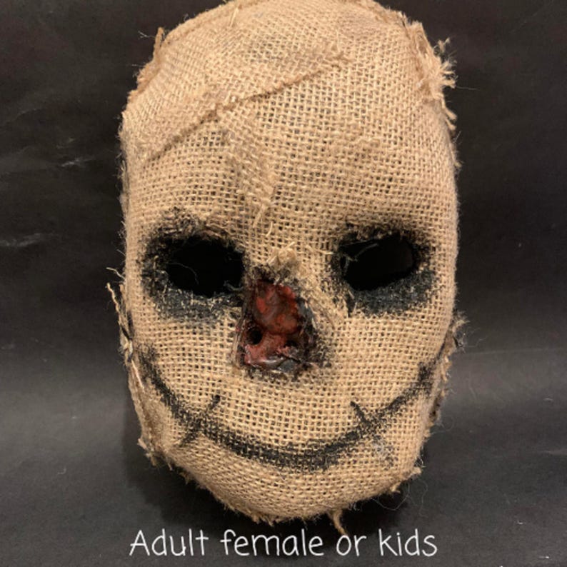 Puede incluir: Una m&aacute;scara de espantap&aacute;jaros de arpillera con costuras, agujeros negros para los ojos y una sonrisa cosida. La nariz est&aacute; pintada de rojo. El texto "Adult female or kids" est&aacute; en la parte inferior de la imagen.