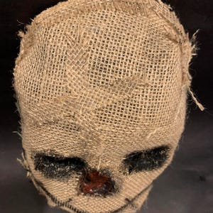 Puede incluir: Una m&aacute;scara de arpillera con rasgos cosidos, que incluyen &aacute;reas negras para los ojos y una sonrisa cosida. La nariz es de un material rojo oscuro y brillante. La m&aacute;scara est&aacute; dise&ntilde;ada para parecerse a un espantap&aacute;jaros.
