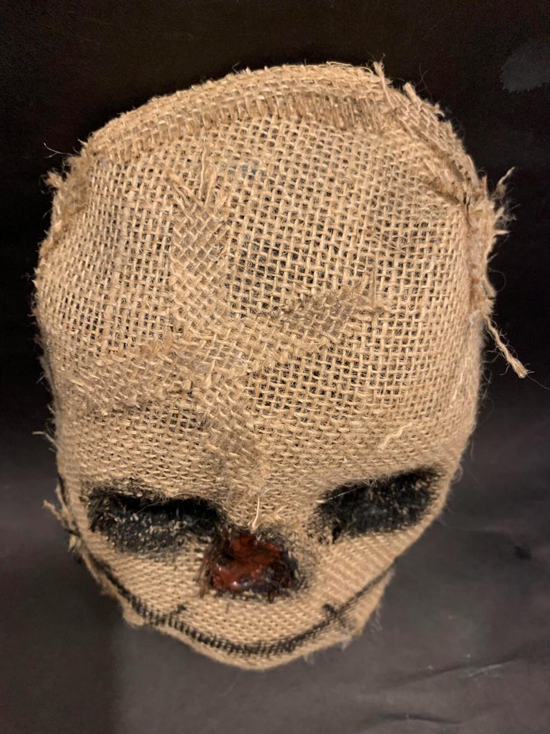 Puede incluir: Una m&aacute;scara de arpillera con rasgos cosidos, que incluyen &aacute;reas negras para los ojos y una sonrisa cosida. La nariz es de un material rojo oscuro y brillante. La m&aacute;scara est&aacute; dise&ntilde;ada para parecerse a un espantap&aacute;jaros.