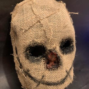 Puede incluir: Una m&aacute;scara de arpillera con costuras y una sonrisa cosida. Los ojos son oscuros y la nariz tiene una apariencia roja y texturizada. La m&aacute;scara est&aacute; dise&ntilde;ada para parecerse a un espantap&aacute;jaros.