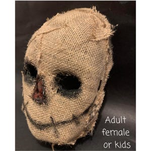 Puede incluir: Una m&aacute;scara de arpillera con grandes agujeros oscuros para los ojos y una sonrisa cosida. El &aacute;rea de la nariz tiene un parche marr&oacute;n rojizo. La m&aacute;scara est&aacute; dise&ntilde;ada para parecerse a un espantap&aacute;jaros. El texto en la imagen dice "Adult female or kids."
