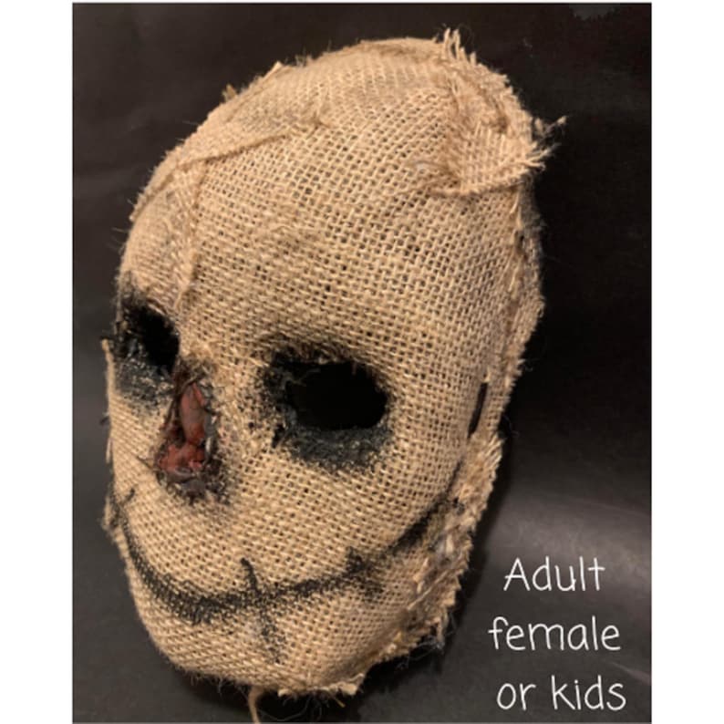 Puede incluir: Una m&aacute;scara de arpillera con grandes agujeros oscuros para los ojos y una sonrisa cosida. El &aacute;rea de la nariz tiene un parche marr&oacute;n rojizo. La m&aacute;scara est&aacute; dise&ntilde;ada para parecerse a un espantap&aacute;jaros. El texto en la imagen dice "Adult female or kids."