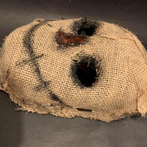 Puede incluir: Una m&aacute;scara de arpillera con grandes agujeros para los ojos y la boca. La m&aacute;scara tiene costuras negras y un parche rojo cerca de la boca. La m&aacute;scara es probablemente para un disfraz de Halloween o una producci&oacute;n teatral.