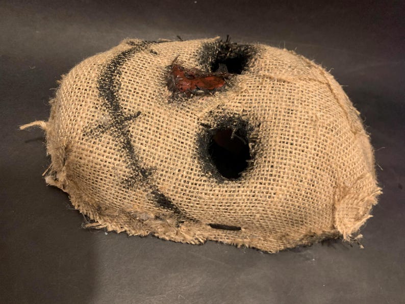 Puede incluir: Una m&aacute;scara de arpillera con grandes agujeros para los ojos y la boca. La m&aacute;scara tiene costuras negras y un parche rojo cerca de la boca. La m&aacute;scara es probablemente para un disfraz de Halloween o una producci&oacute;n teatral.